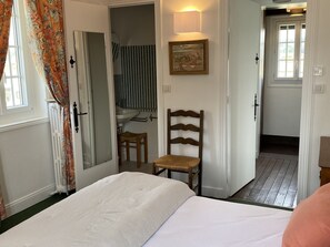 A bedroom with a bed, a wooden chair, a small table, a mirror, and a painting on the wall.