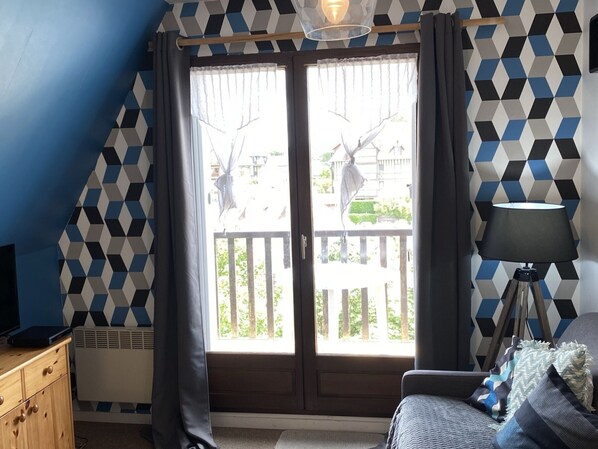 Una habitación con pared de diseño geométrico, puerta de cristal corredera que da a un balcón, escritorio con una computadora portátil, lámpara y sofá con cojines.