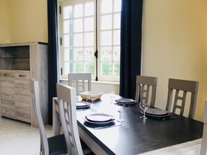A dining area with a table set for four, chairs, a window with curtains, and a wooden cabinet. - Ground Floor Renovated 2-Room Apartment Near Cabourg Center with Private Garden (Cabourg)