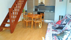 A compact living space with a wooden dining table and chairs, a sofa with patterned upholstery, a kitchen area with modern appliances, and a wooden staircase. - Duplex 3-Room Apartment Near Beach in Cabourg - Sleeps 6, Parking Included (Le Home Varaville)