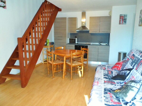 Um espaço de estar compacto com uma mesa de jantar de madeira e cadeiras, um sofá com estofamento estampado, uma área de cozinha com eletrodomésticos modernos e uma escada de madeira.