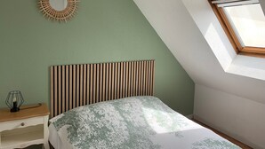 A bedroom with a sloped ceiling, a bed with a patterned bedspread, a bedside table with a lamp, and a wall-mounted mirror. - Duplex 3-Room Apartment Near Beach in Cabourg - Sleeps 6, Parking Included (Le Home Varaville)