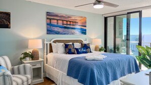 A bedroom with a bed, a chair, a nightstand, a lamp, a painting, and a view of the ocean through a window. - Family Beach Condo - Steps To The Sand Or Pool (St. Augustine)