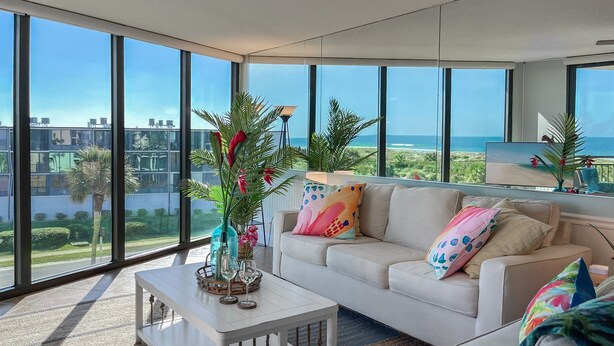 A modern living room with a large window overlooking a coastal view, a white sofa with colorful pillows, and a coffee table with decorative items. - Family Beach Condo - Steps To The Sand Or Pool (St. Augustine)