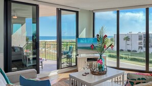 A spacious living room with a large TV, comfortable seating, a coffee table, and a balcony with a view of the ocean. - Family Beach Condo - Steps To The Sand Or Pool (St. Augustine)