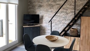Une cuisine moderne avec un mur de crédence en pierre, un coin repas avec une table ronde et des chaises, un téléviseur posé sur un meuble, et un escalier avec une rampe en métal.