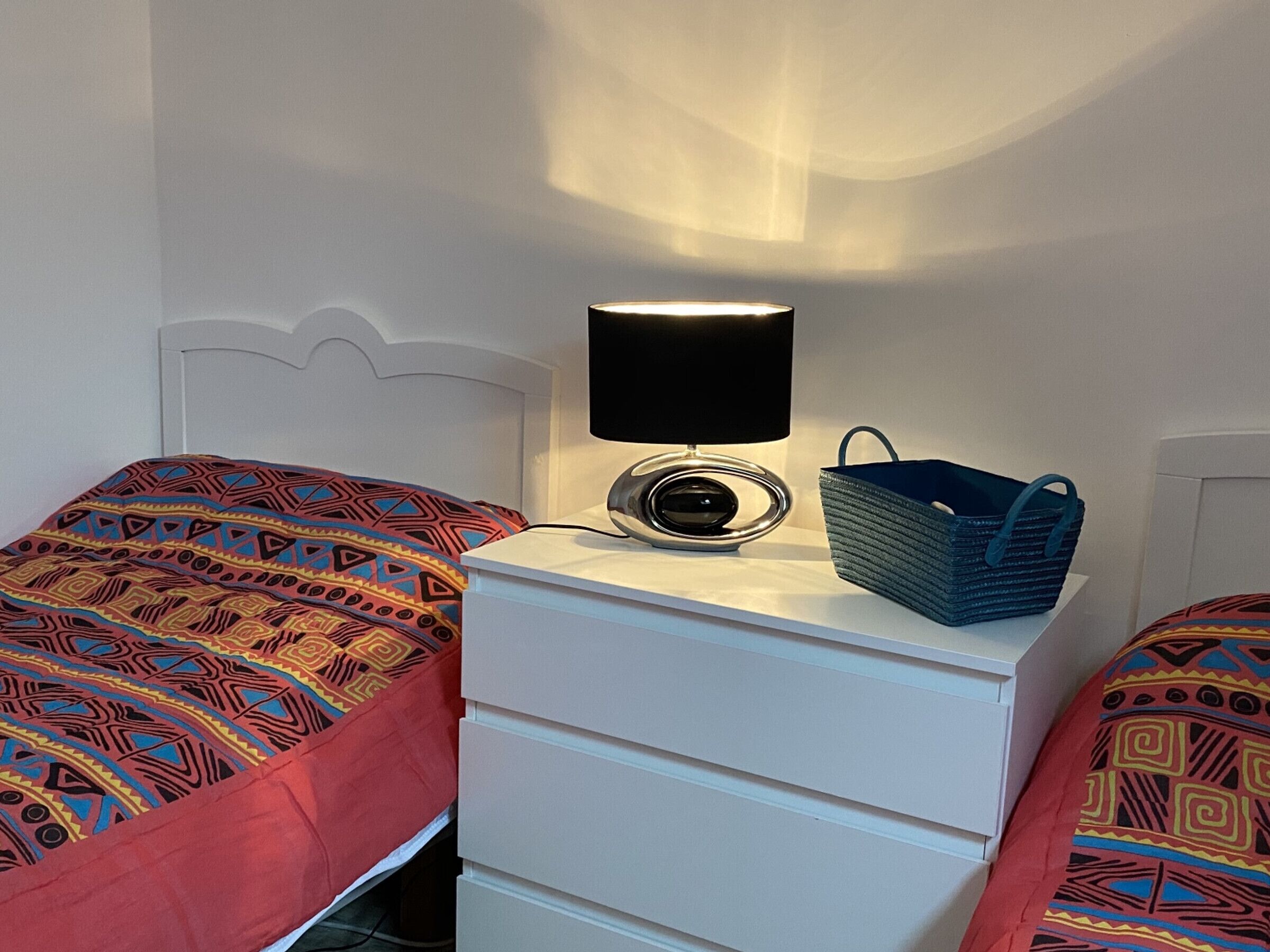 En seng med et mønstret tæppe, et natbord med en lampe og en blå vævet taske, samt en hvid sengegavl.