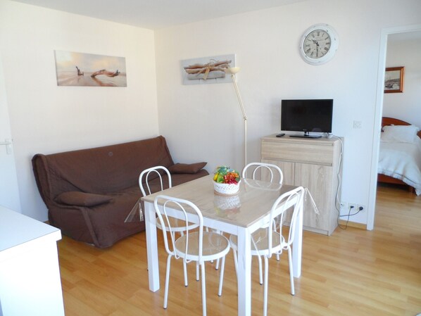 A cozy living room with a brown sofa, a dining table with white chairs, a TV on a wooden cabinet, and artwork on the walls.
