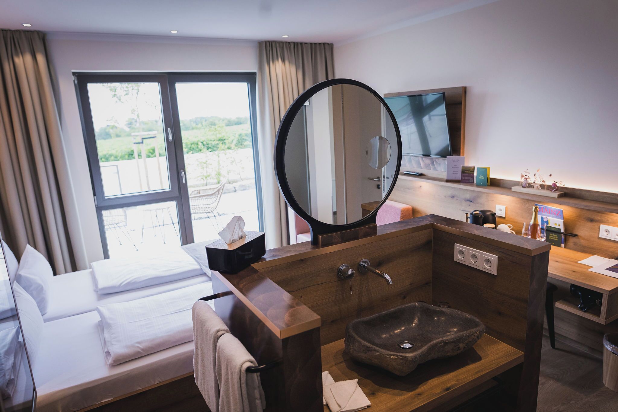 A hotel room with a bed, a large mirror, a sink with a stone basin, a television, and a window with curtains.