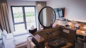 A hotel room with a bed, a large mirror, a sink with a stone basin, a television, and a window with curtains.