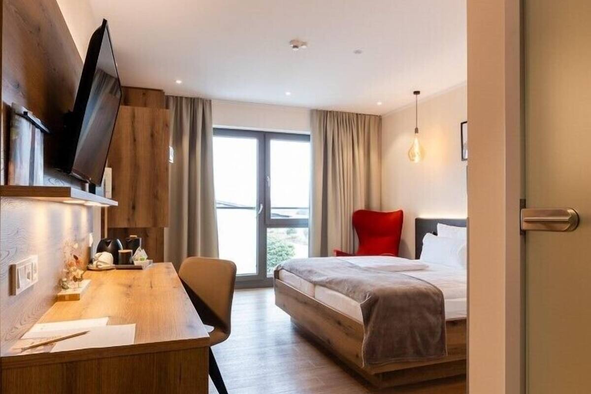 A modern hotel room with a wooden desk, a bed with red pillows, and a large window offering a view.