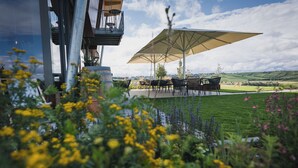 Un espacio al aire libre con mesas y sillas bajo un gran paraguas, rodeado de abundante follaje y flores.