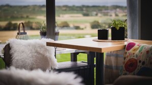 Un cuarto acogedor con vistas a un paisaje verde a través de amplios ventanales, una mesa de madera con dos plantas en macetas, un almohadón blanco y mullido, y un cojín colorido.