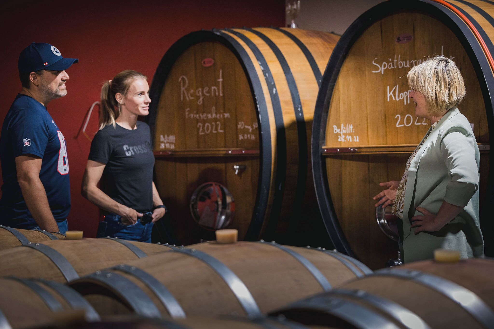 Three people standing in front of wooden barrels in a cellar.