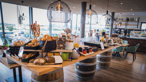 Mostrador de panadería con diversos productos horneados y pasteles, barriles de madera como taburetes y lámparas colgantes.