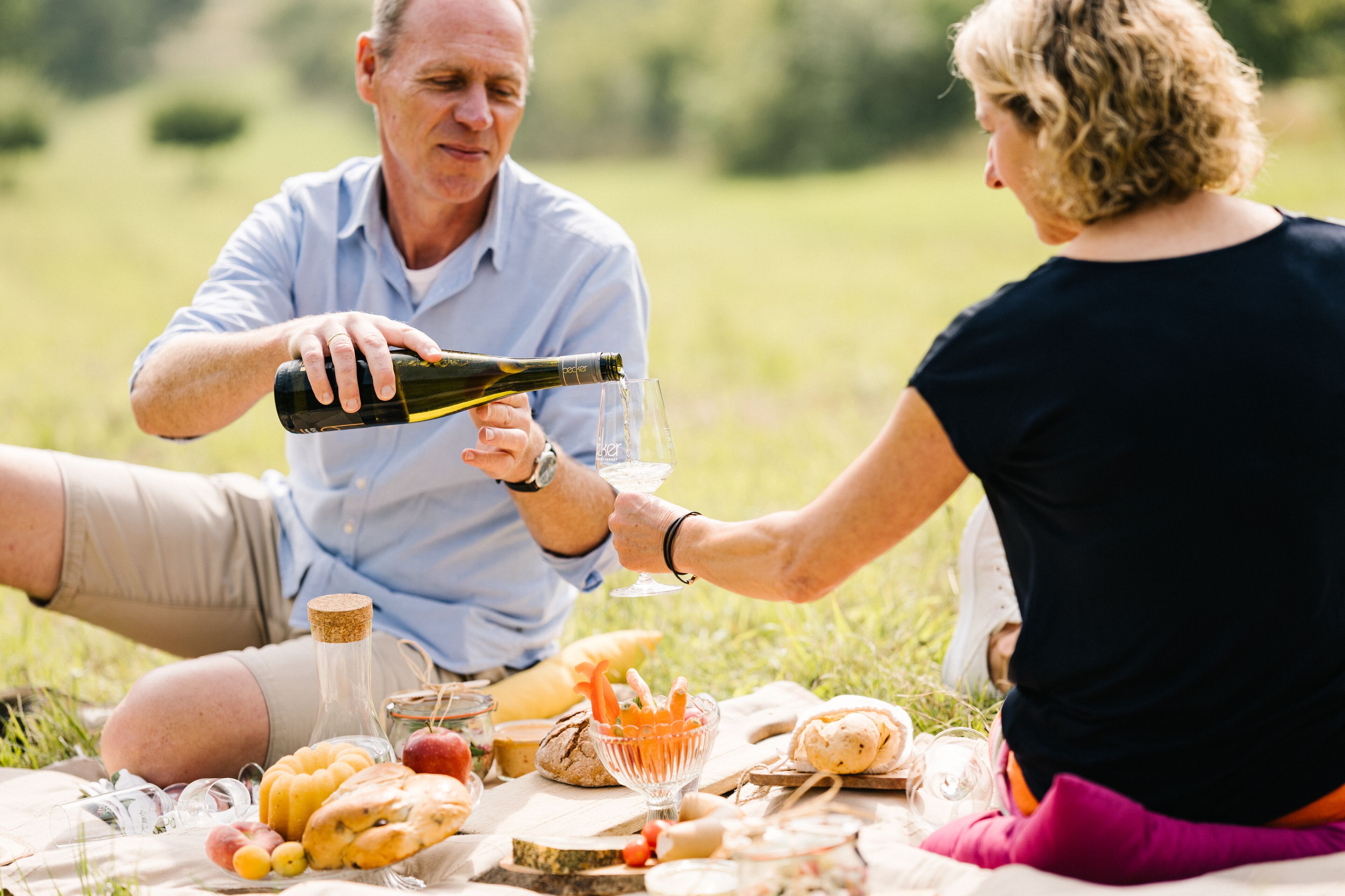 A picnic with a bottle of wine, glasses, and various food items on a blanket in a grassy area.