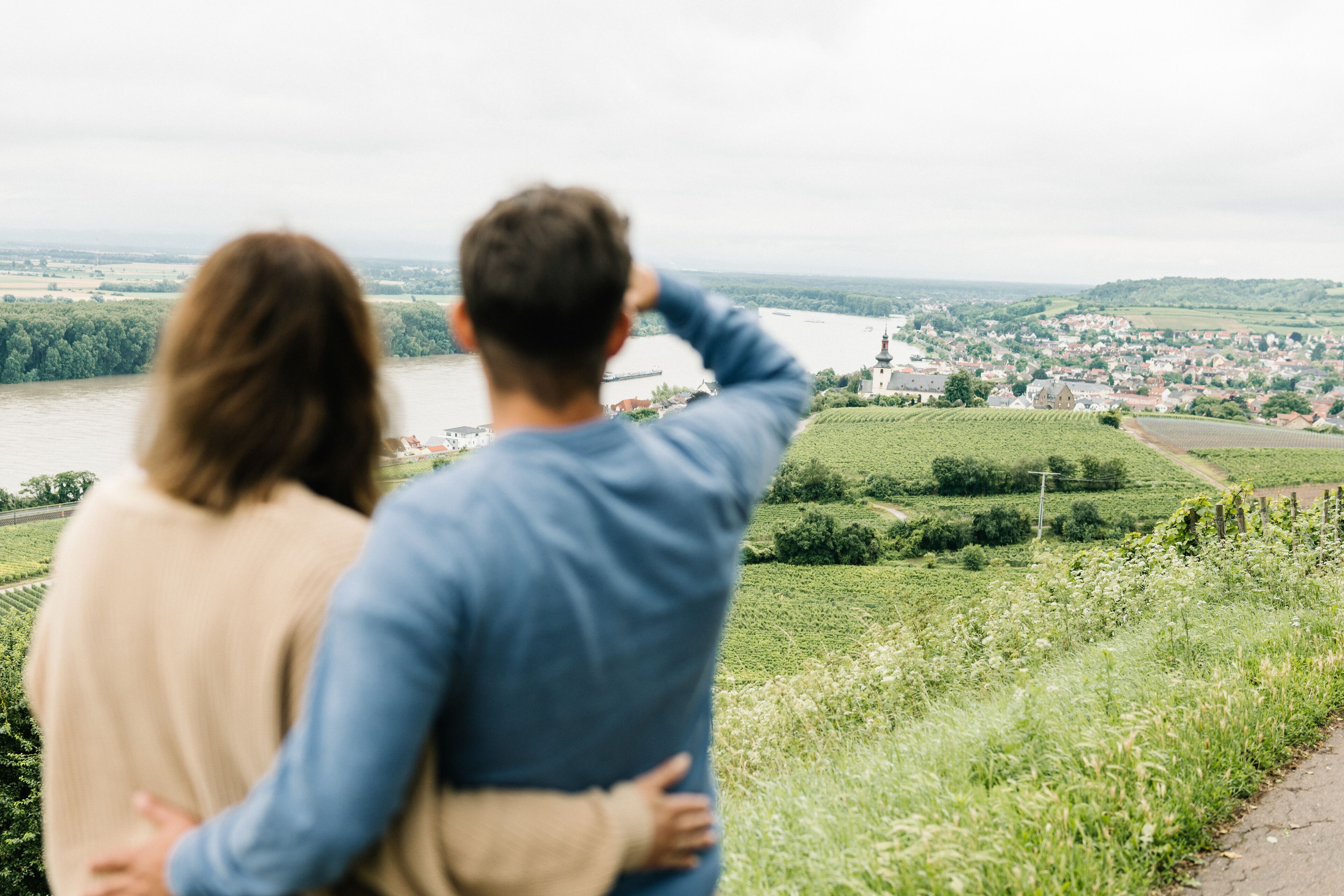 A couple standing on a grassy hill overlooking a river and a town.