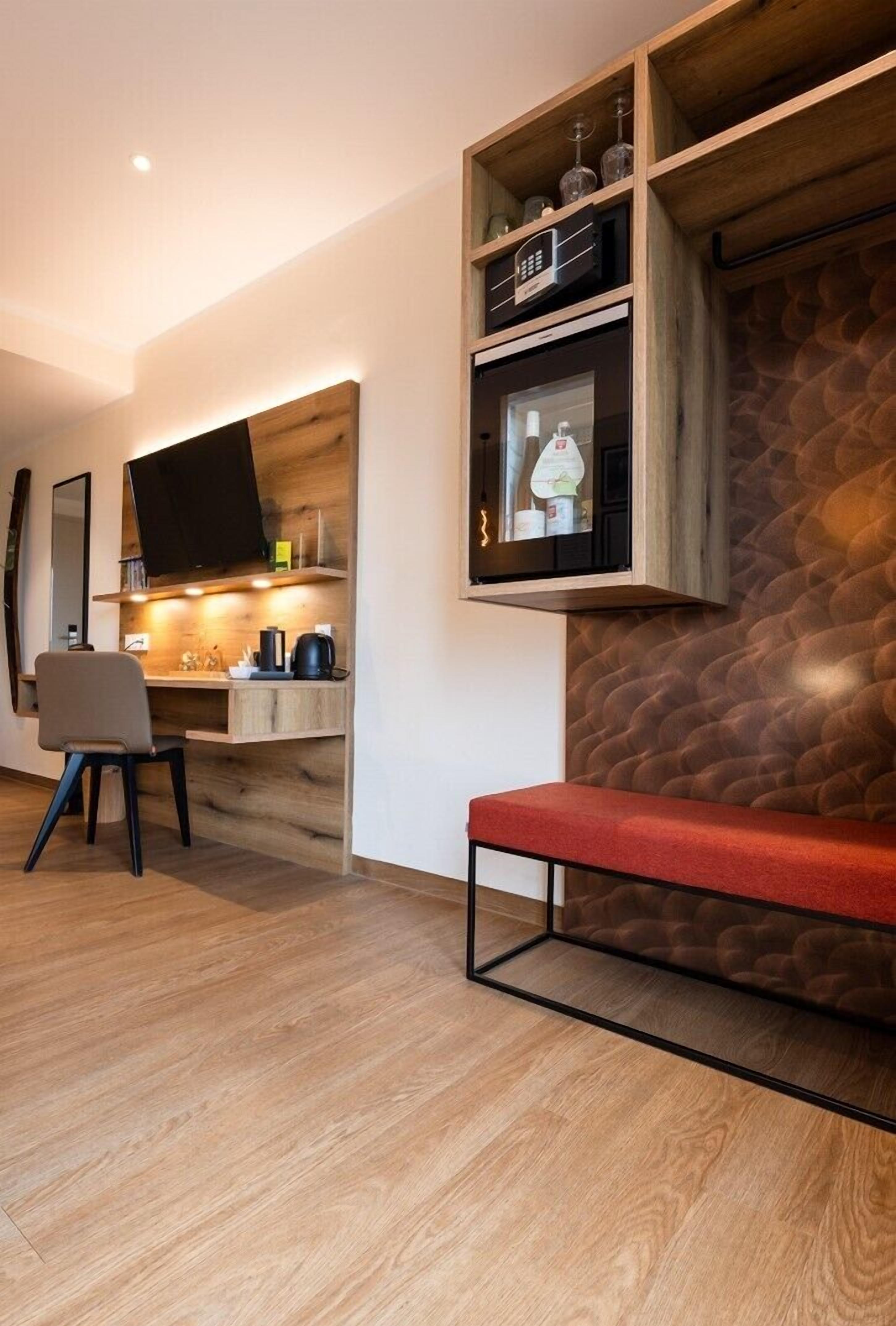 A modern living room with a wooden floor, a red bench, a TV mounted on a wooden shelf, and a kitchen area with a stove and sink.