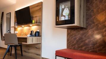 A modern living room with a wooden floor, a red bench, a TV mounted on a wooden shelf, and a kitchen area with a stove and sink.