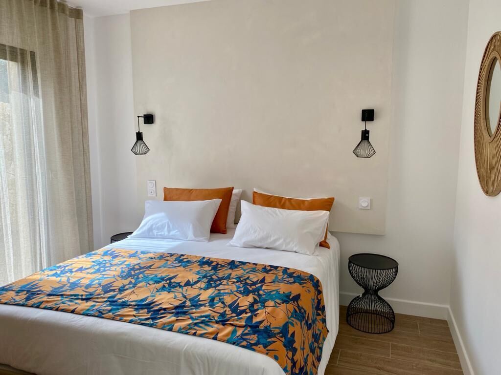 Een net opgemaakt bed met een bloemenbedekking, twee wandlampen en een ronde spiegel aan de rechtermuur.
