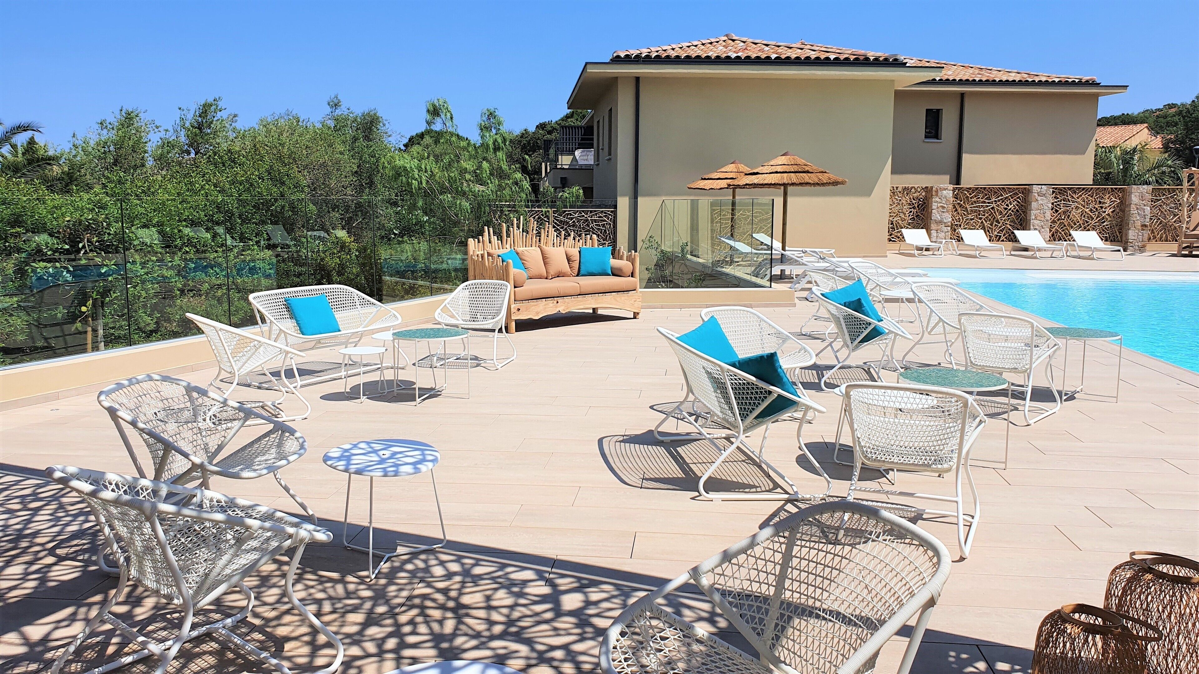 A poolside area with lounge chairs and a sofa, surrounded by a fence and trees.