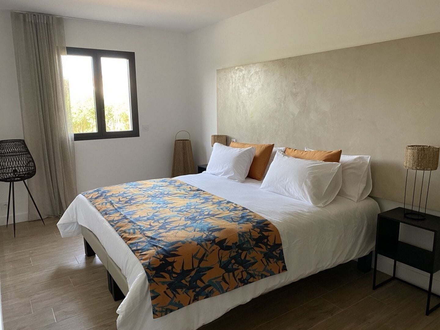 A neatly made bed with a patterned bedspread, a bedside lamp, and a chair in a modern room with a large window and a plain wall.