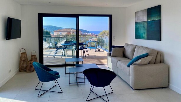 Ein modernes Wohnzimmer mit einer Couch, zwei Sesseln, einem Fernseher und einem Balkon mit Gartenmöbeln.