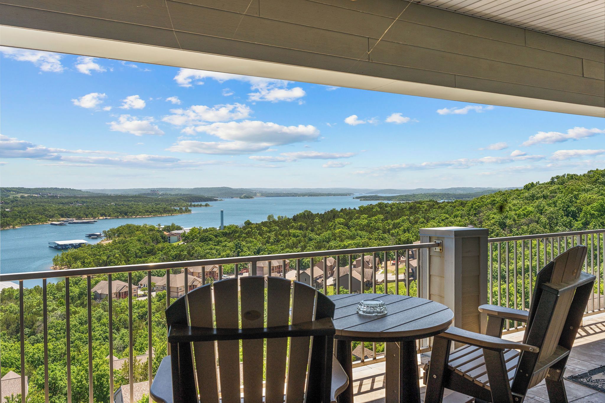 A balcony with two Adirondack chairs and a small table, overlooking a river and a forested area.