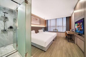 A modern hotel room with a glass-enclosed shower, a large bed with two pillows, a desk with a chair, and a television on the wall.