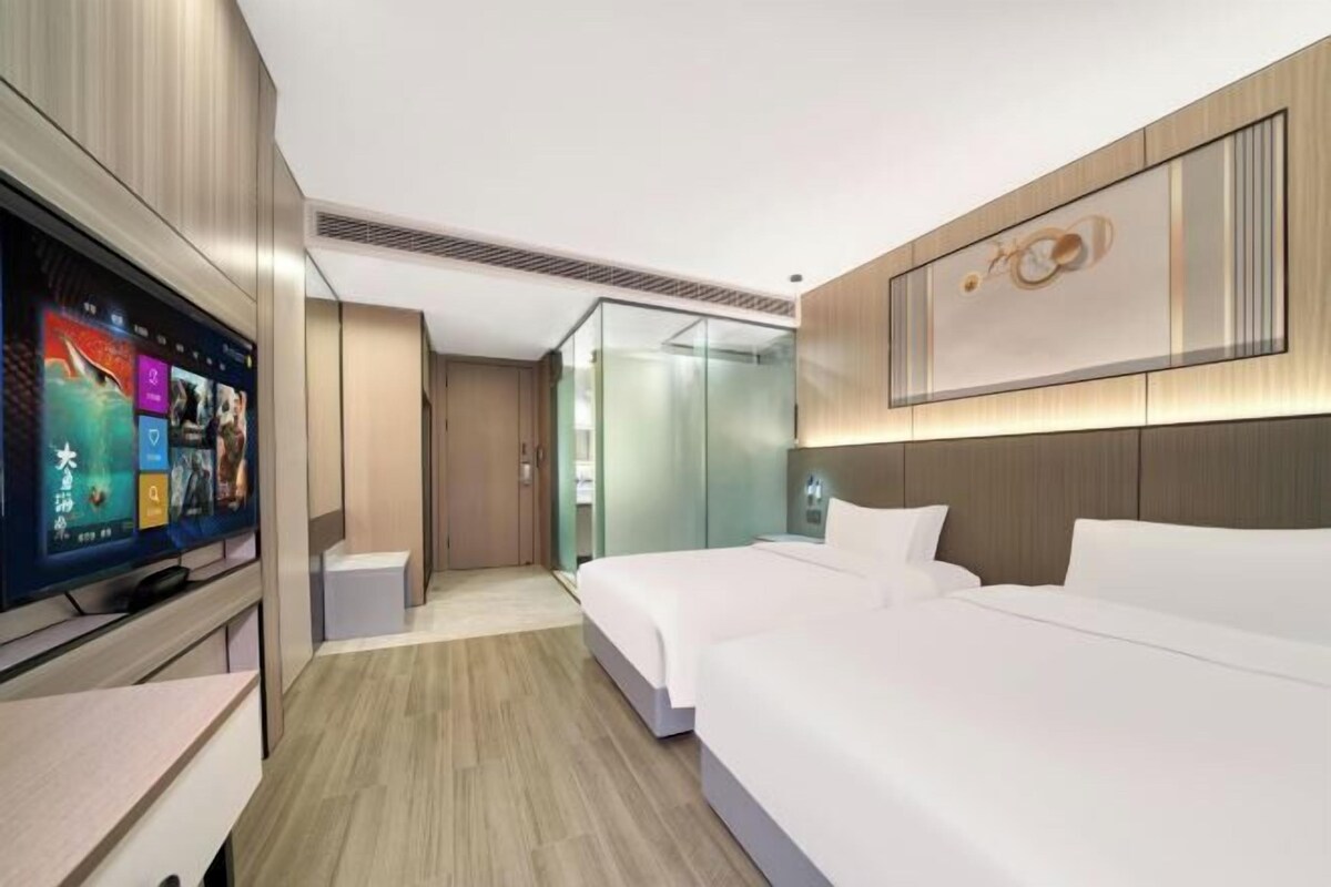 A modern hotel room with a large flat-screen TV, two beds, a glass partition, and a wooden headboard.