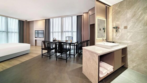 A modern hotel room with a large bed, a dining area with a table and chairs, a bathroom with a large mirror and sink, and a view of the city through large windows. - Four Seasons Qingteng Hotel (Outlet Airport) (Ningbo)