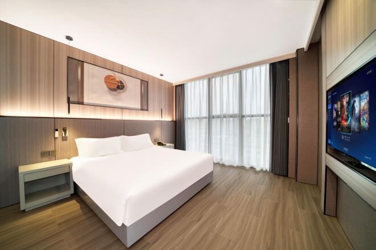 A modern hotel room with a large bed, a flat-screen TV, and a large window with curtains.