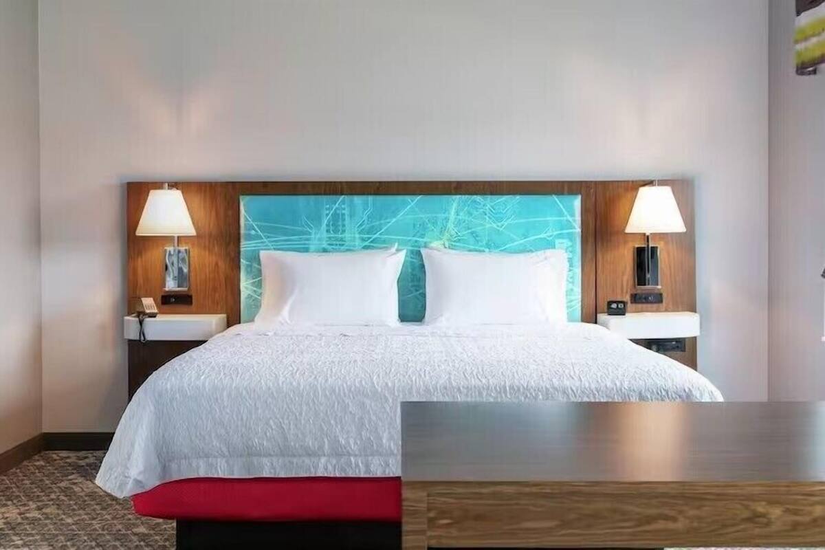 A hotel room with a large bed, two bedside tables with lamps, a wooden headboard with a geometric design, and a red bench at the foot of the bed.