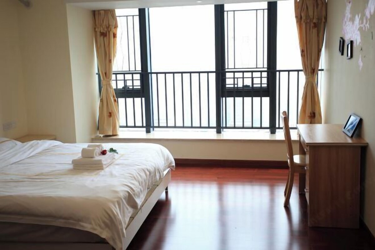 A hotel room with a bed, a desk, and a view of the outside through large windows.