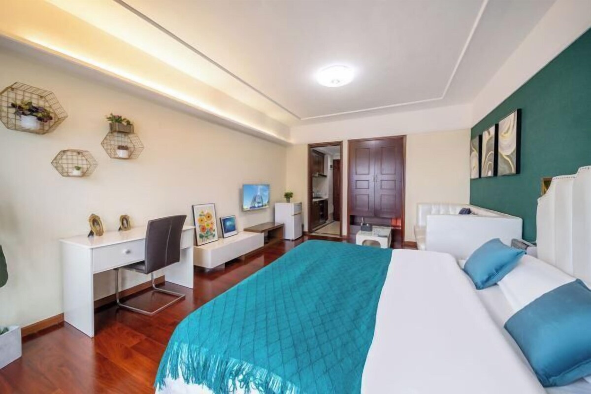 A modern bedroom with a bed, desk, and chair, a small kitchenette, and a bathroom.