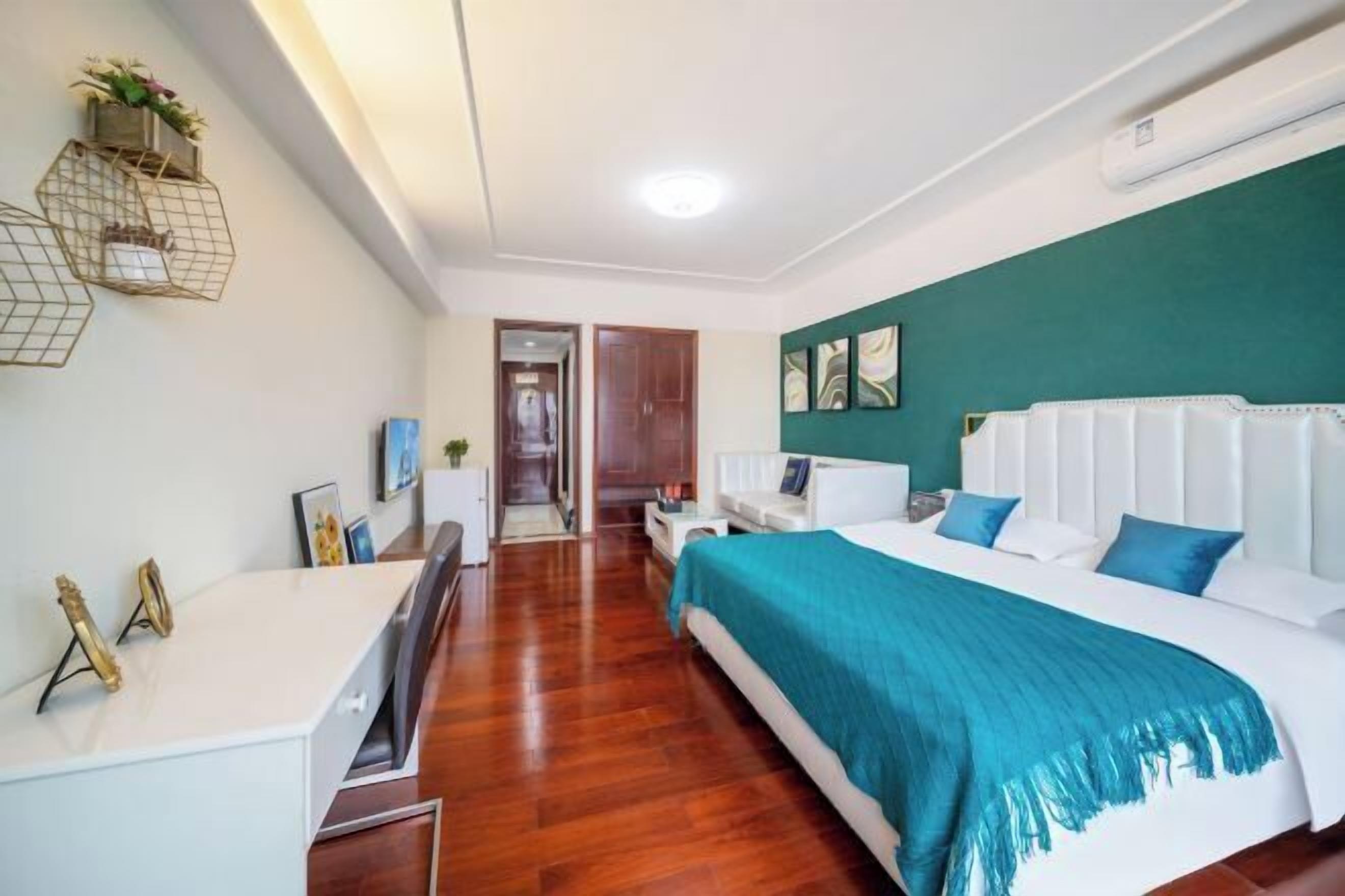 A hotel room with a wooden floor, a bed with a teal bedspread, a white desk, and a white chair.