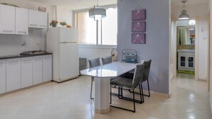 Une cuisine moderne avec des meubles blancs, une table à manger et des chaises. On y trouve une fenêtre avec des rideaux, un luminaire suspendu et une petite étagère ornée d’objets décoratifs.