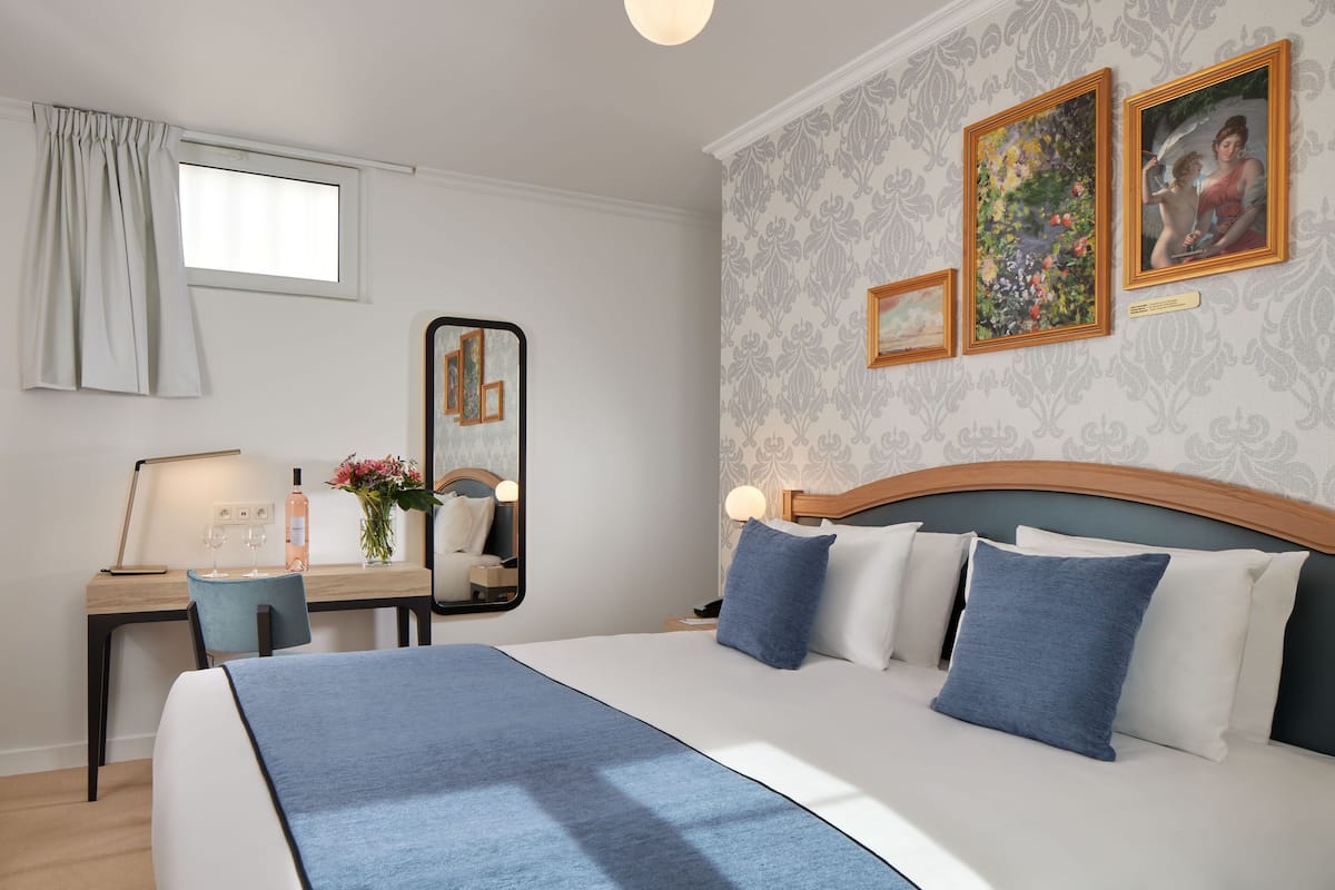 A hotel room with a large bed, a desk with a chair, a mirror, and two paintings on the wall.