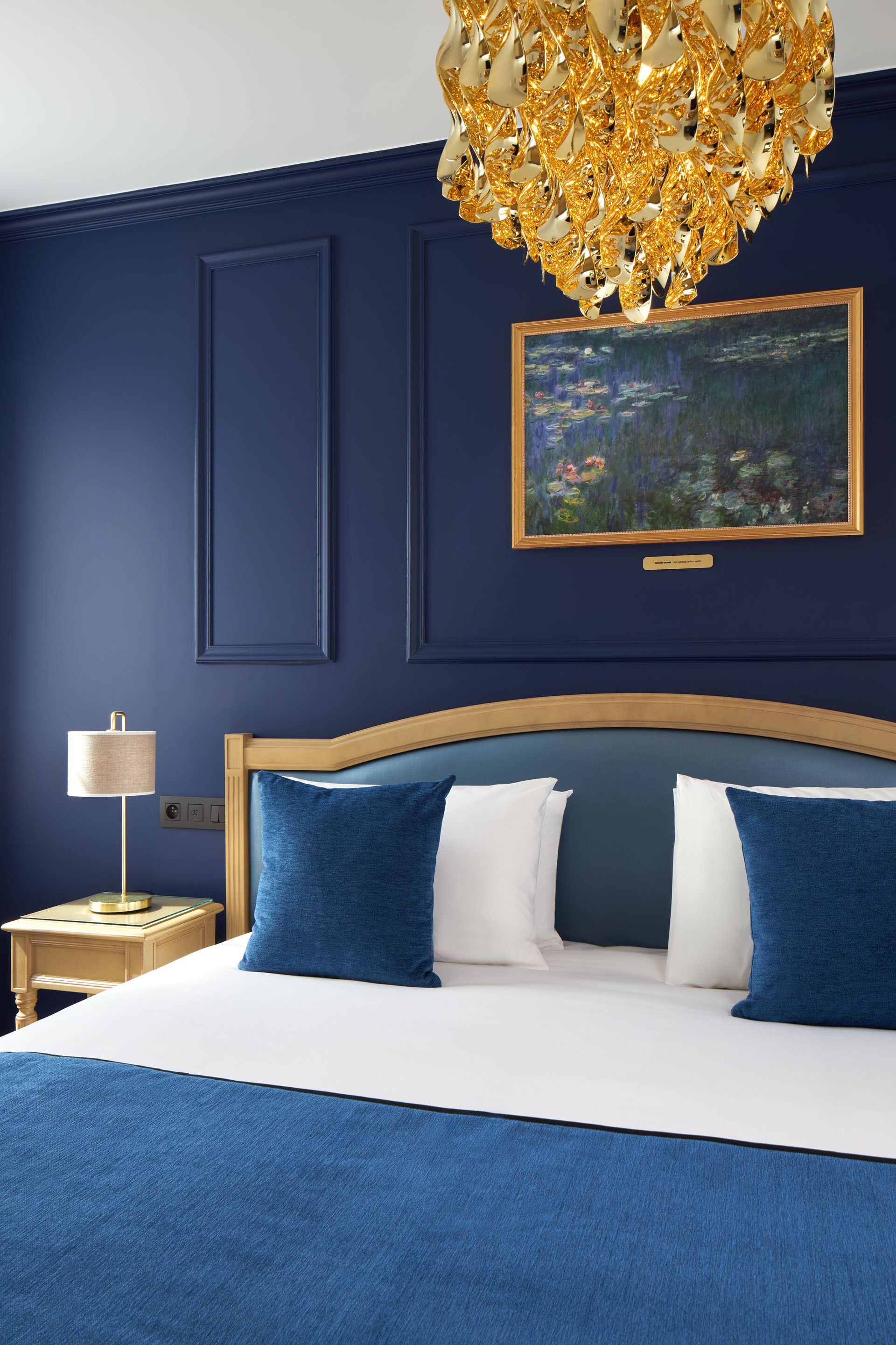 Quarto com cama grande, cabeceira de madeira, esquema de cores azul e branco, lustre dourado e quadro pendurado na parede.