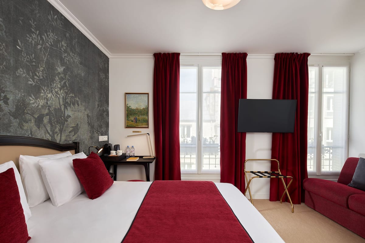 A hotel room with a large bed, red curtains, a desk with a lamp, a TV, and a red sofa.