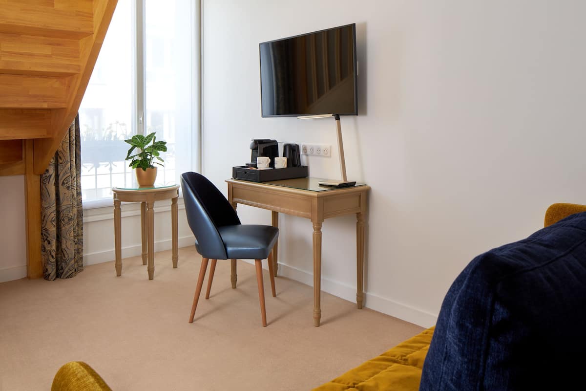 A modern hotel room with a wooden desk, a chair, a television, a coffee maker, and a potted plant.