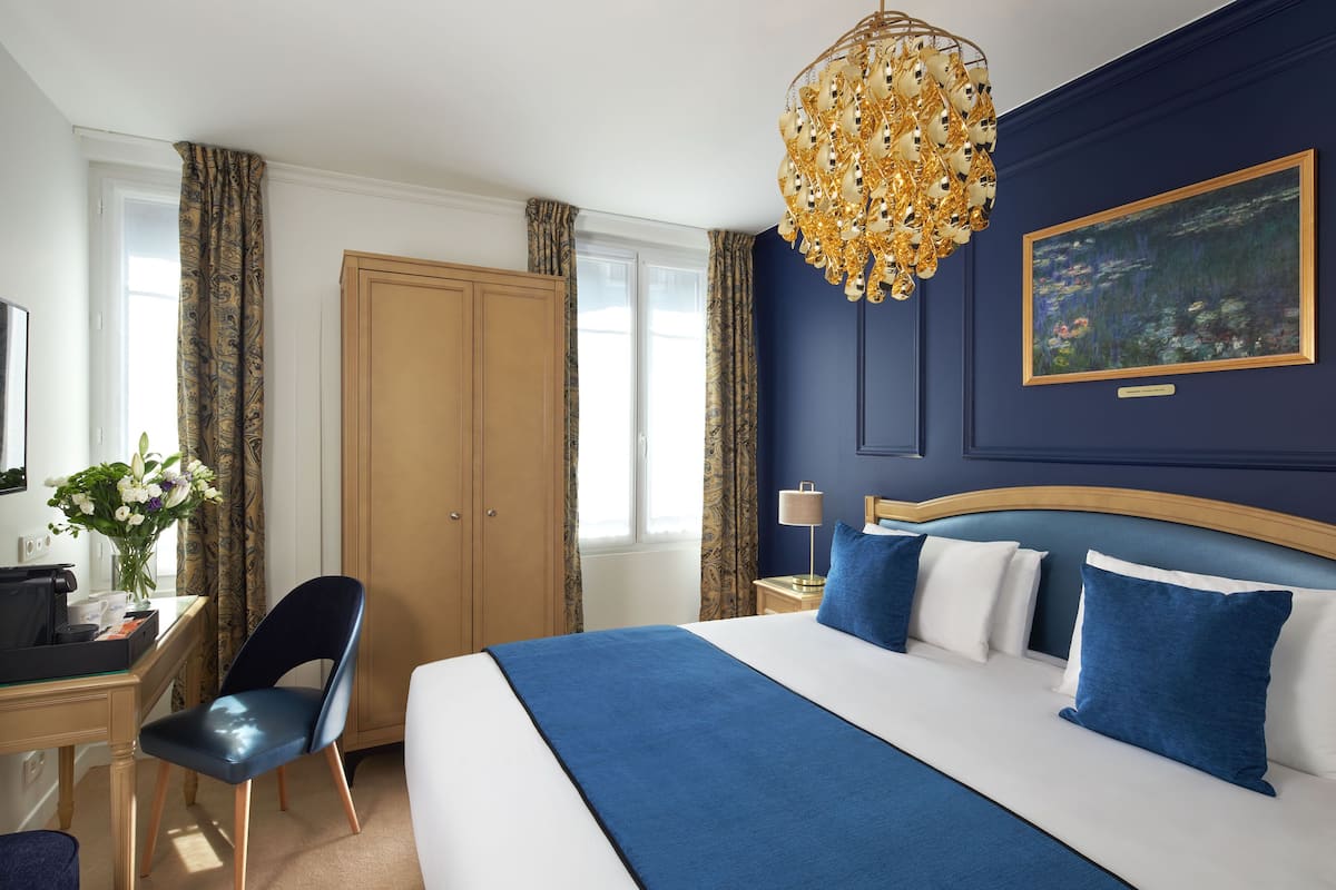A hotel room with a large bed, a desk with a computer, a chair, a desk lamp, and a painting on the wall.