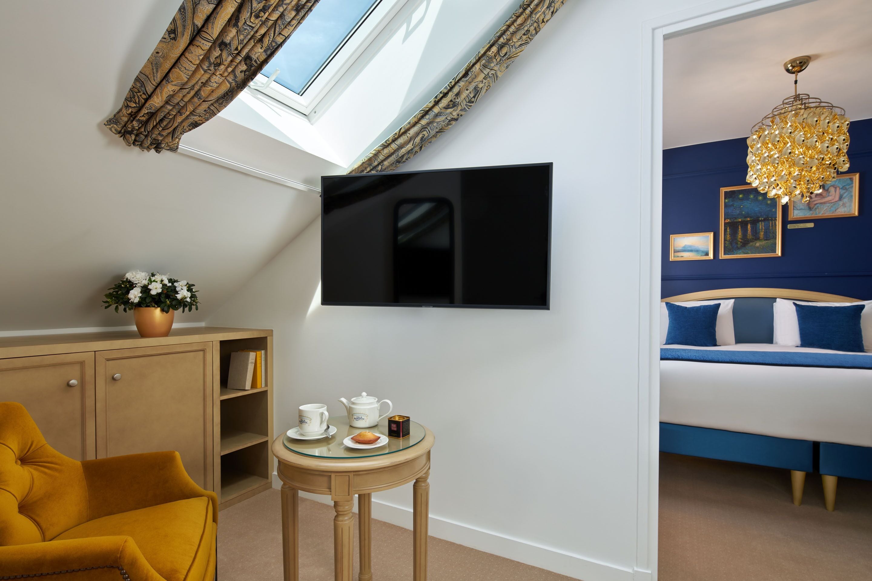 Une chambre avec un lit, un téléviseur fixé au mur, une petite table avec une théière et une tasse, et une fenêtre avec des rideaux.