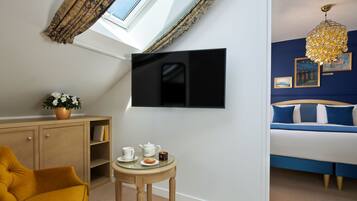 Une chambre avec un lit, un téléviseur fixé au mur, une petite table avec une théière et une tasse, et une fenêtre avec des rideaux.