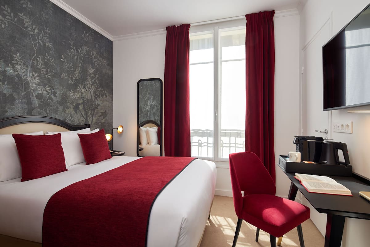 A hotel room with a large bed, red curtains, a desk with a coffee maker, and a TV on the wall.