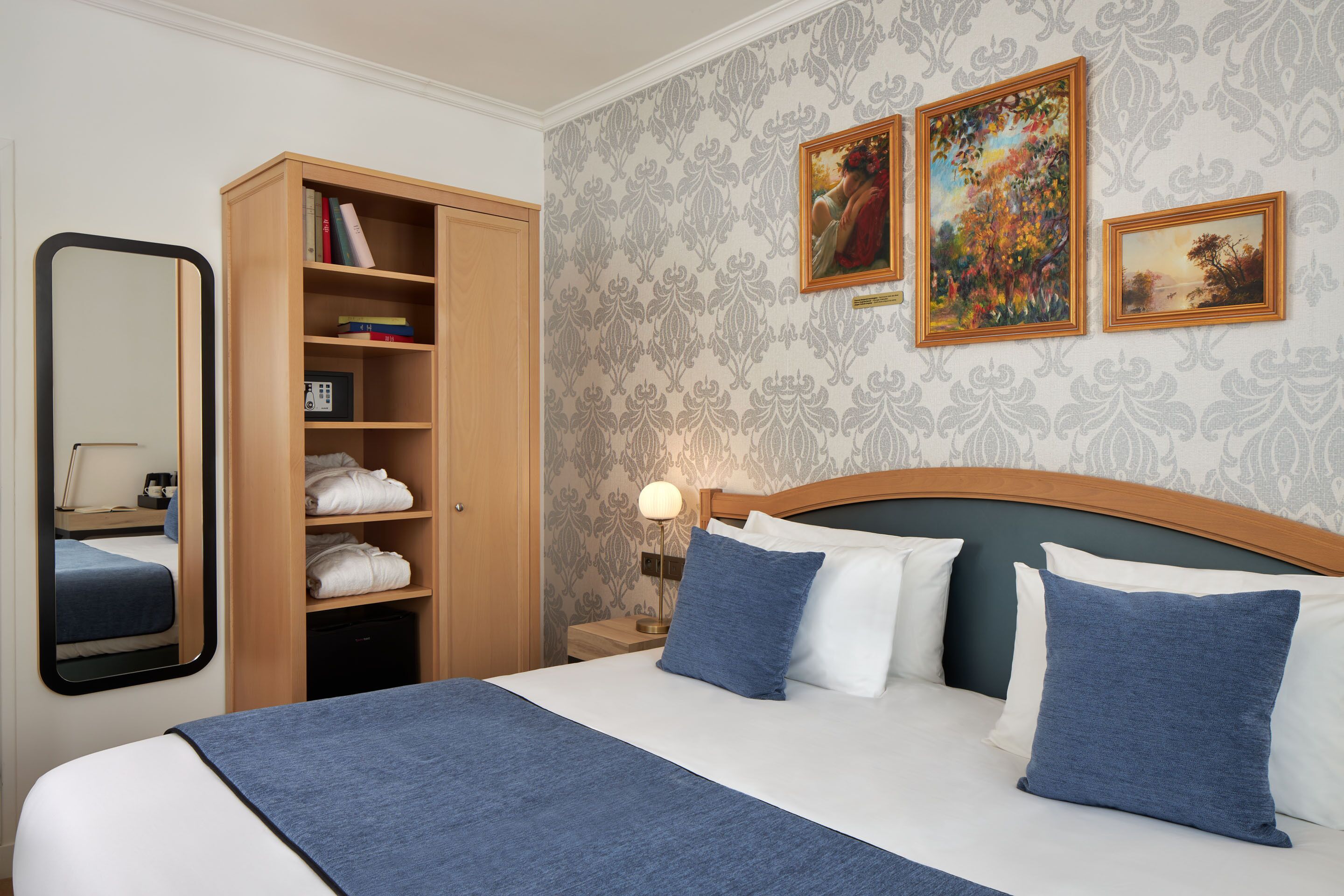 A neatly arranged hotel room with a bed, a wardrobe, a bedside table with a lamp, and two framed artworks on the wall.