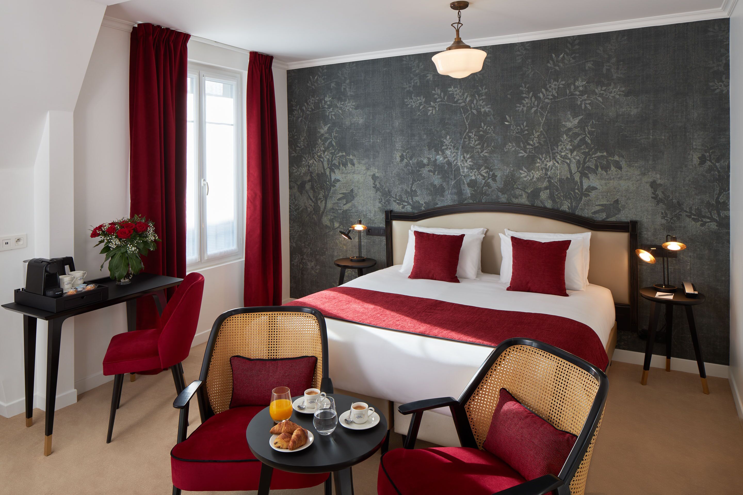 Habitació d'hotel amb un llit gran, detalls vermells, zona de menjador amb cadires i taula, i una petita taula auxiliar amb una làmpada.