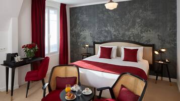 Habitació d'hotel amb un llit gran, detalls vermells, zona de menjador amb cadires i taula, i una petita taula auxiliar amb una làmpada.