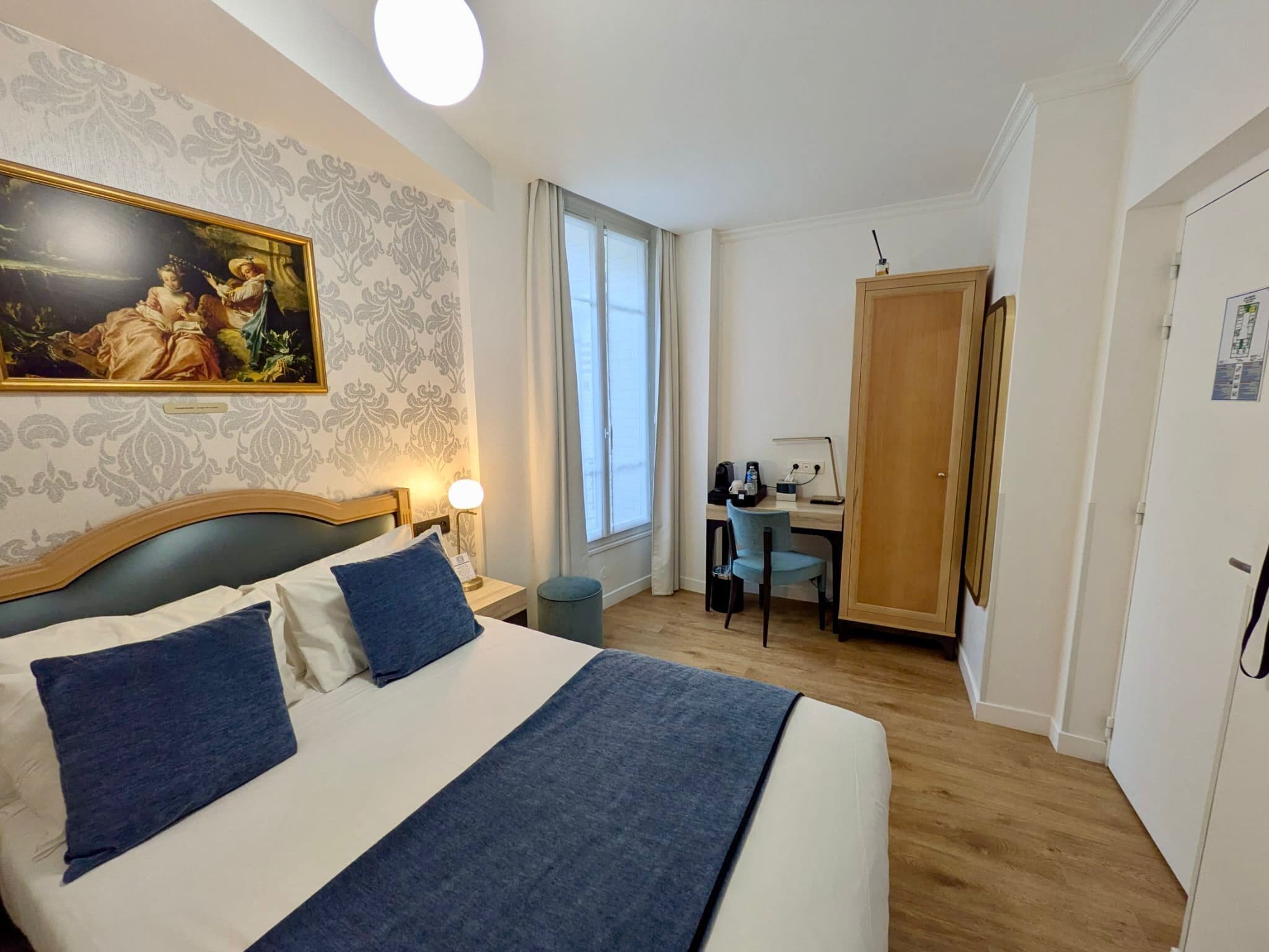 A hotel room with a bed, a desk, a chair, a lamp, and a painting on the wall.