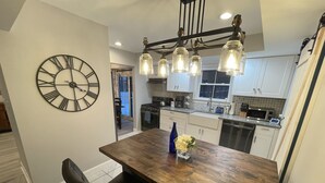 A modern kitchen with a wooden dining table, a large wall clock, and pendant lighting.